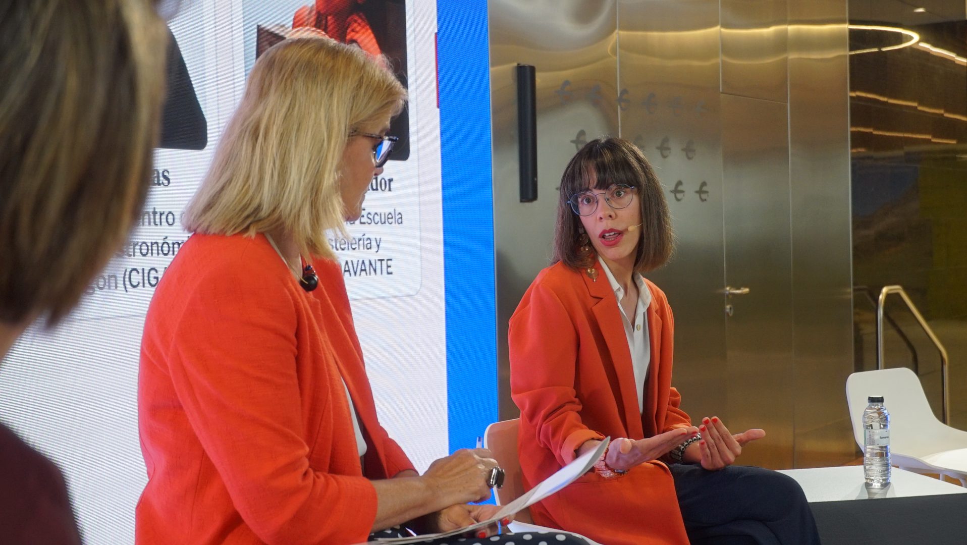 Raquel y Anabel sentadas en sillas altas durante el evento con una filmina digital de fondo.